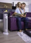 Purificatore d'aria Ionic-CARE in ambienti chiusi s fialovou pohovkou, na které sedí otec a čte knížku své malé dceři, colore bianco perlato