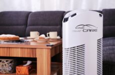 Purificatore d'aria Ionic-CARE in dettaglio u konferenčního stolku s kávou a koláčem, vedle tmavě šedé pohovky, colore bianco perlato